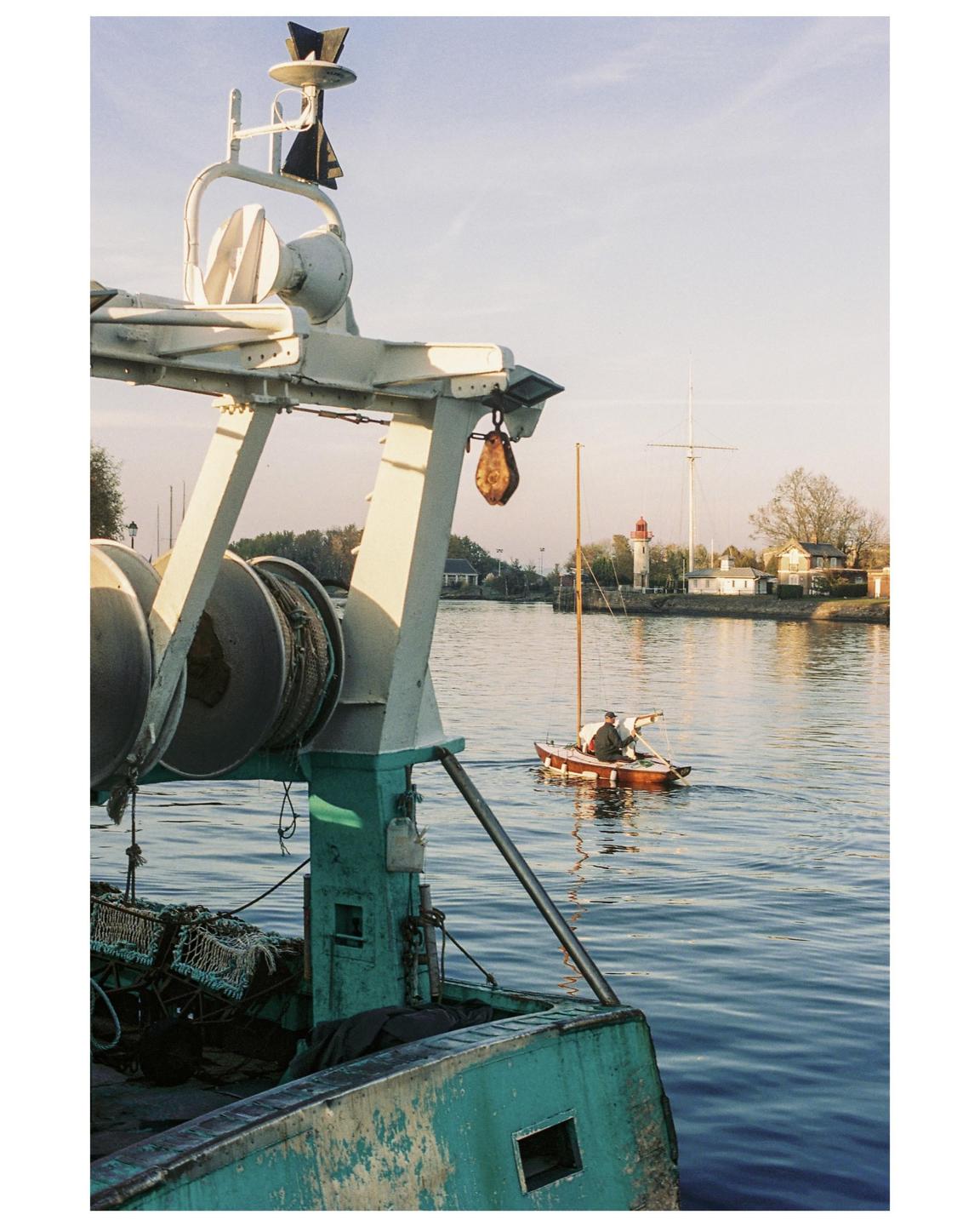 A man rows a small boat past a fishing vessel in a harbor with a lighthouse in the background.
