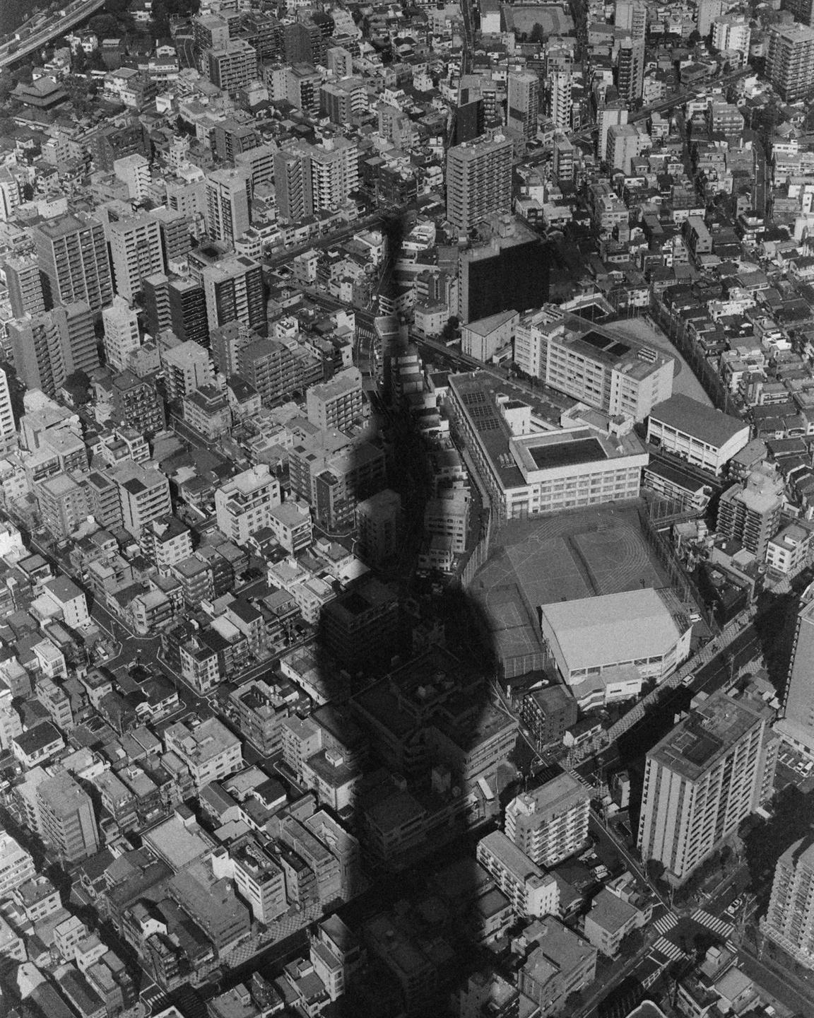 An aerial view shows the shadow of a tall tower cast over a dense urban landscape.