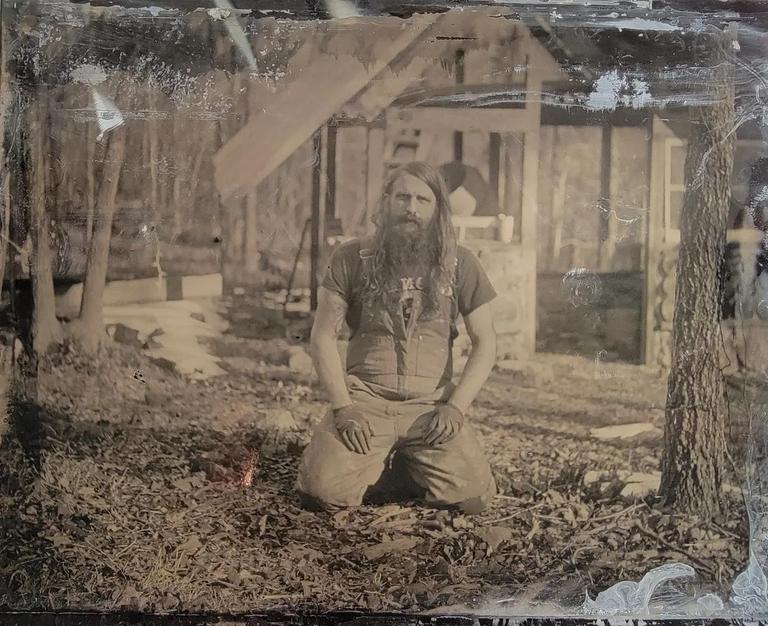 A man with a long beard and hair kneels on the ground in front of a wooden structure.