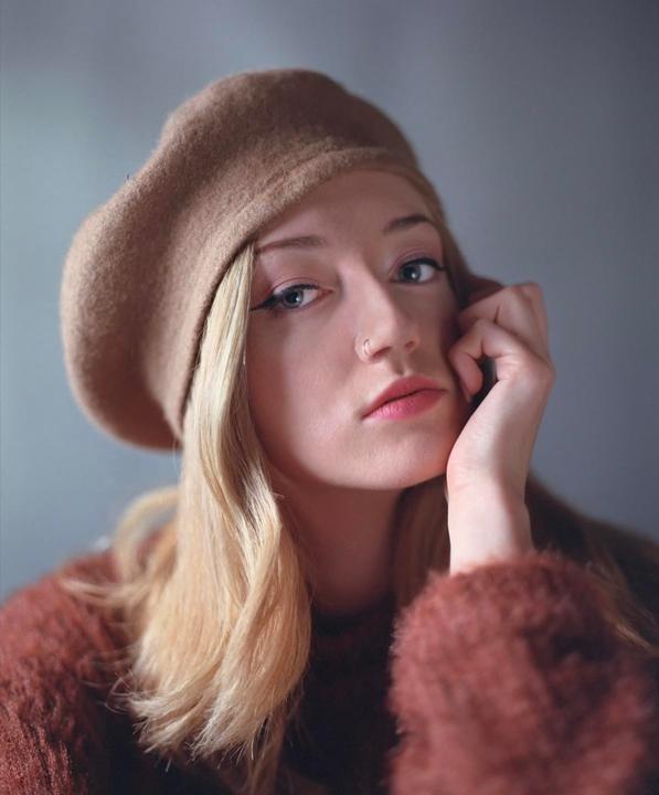 A woman wearing a beret and a fuzzy sweater poses for a portrait with her hand resting on her chin.