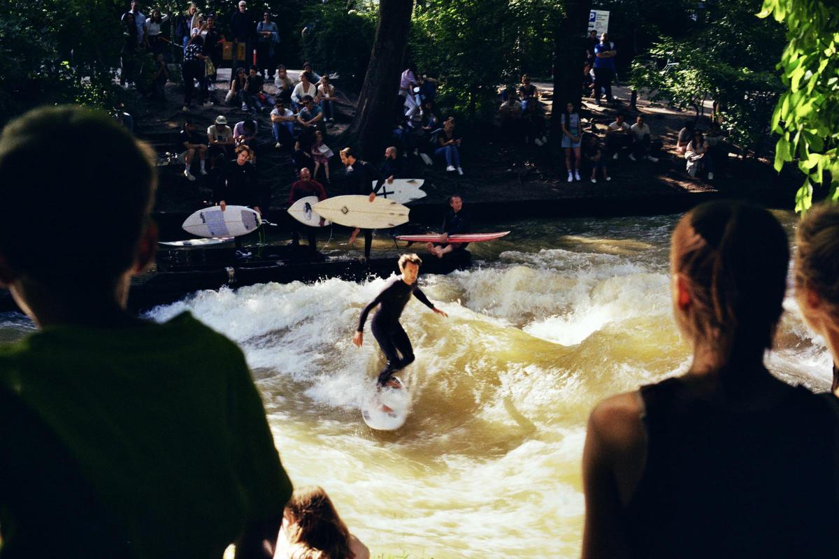A surfer in a wetsuit rides a wave on a river while people watch from the bank.