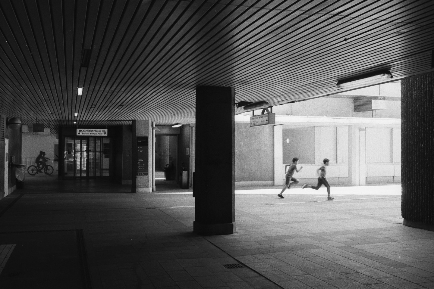 Two boys run through a covered walkway while another boy on a bicycle is visible in the background.