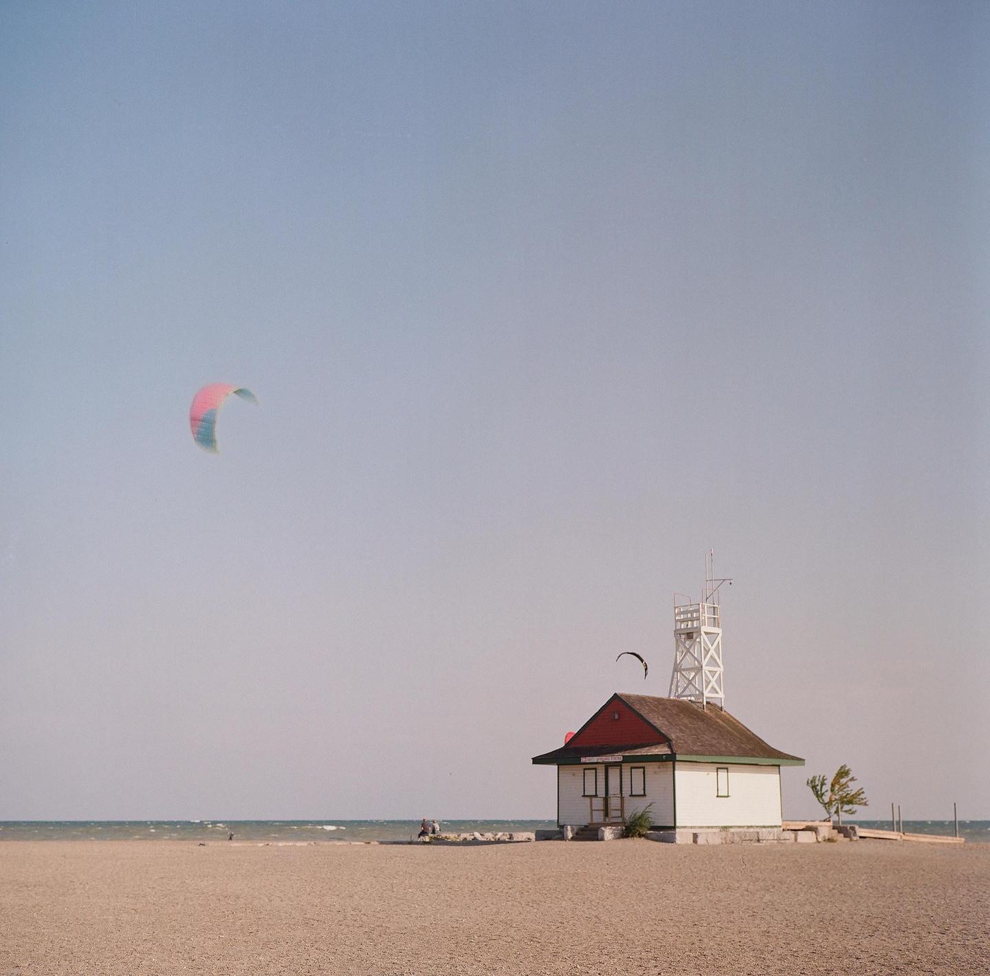 A lifeguard station with a red roof stands on a sandy beach with a kite in the sky and the ocean in the background.