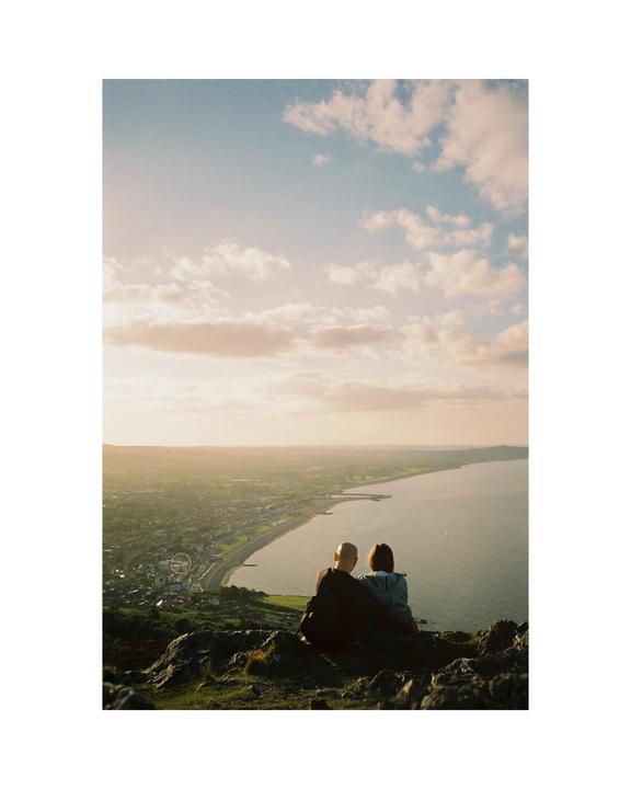 A couple sits on a hill overlooking a coastal town and the sea at sunset.