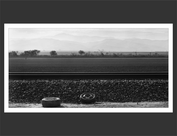 Two discarded tires rest on the gravel next to a railroad track, with a field and hazy mountains in the background.