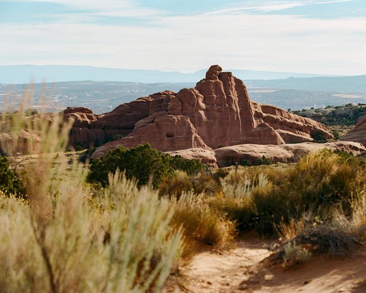 A large red rock formation rises above a desert landscape with sparse vegetation under a blue sky.