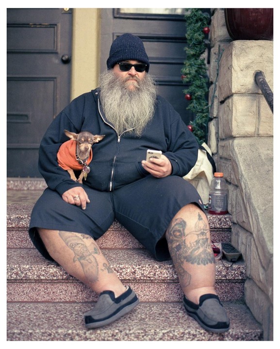 A man with a long beard and sunglasses sits on steps with a small dog in an orange sweater.
