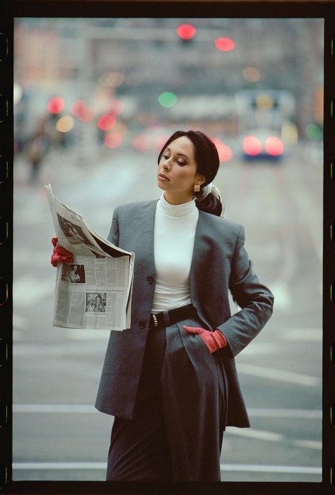 A woman in a gray suit and red gloves holds a newspaper on a city street.