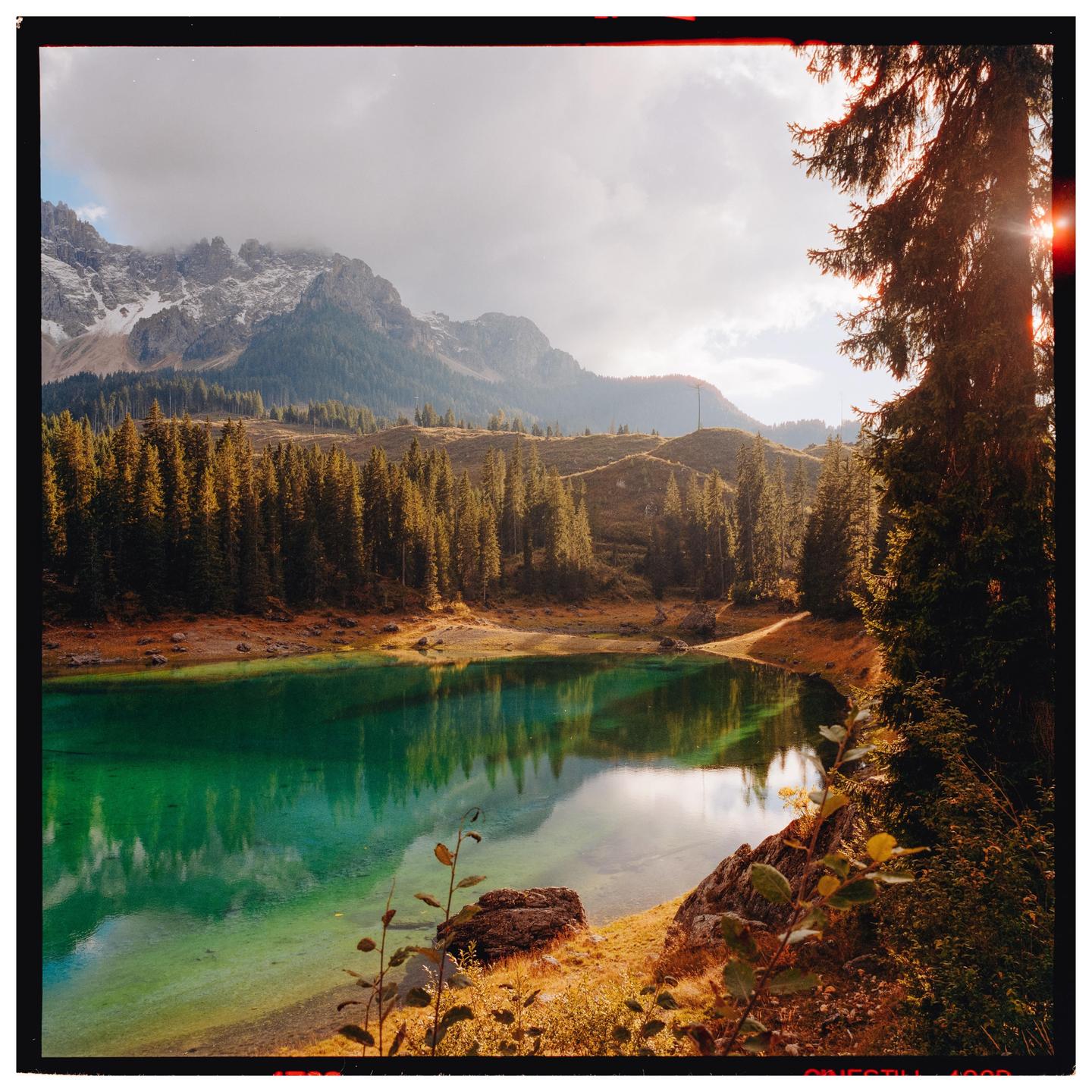 A vibrant green lake is surrounded by a forest of pine trees and mountains under a cloudy sky.