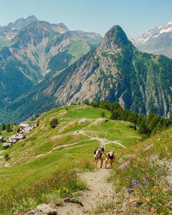 Two people hike up a winding dirt path on a grassy mountain with a village and snow-capped peaks in the distance.