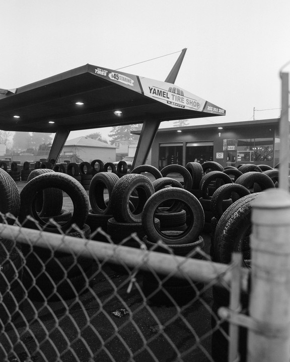 A tire shop with many tires stacked behind a chain link fence is shown on a hazy day.