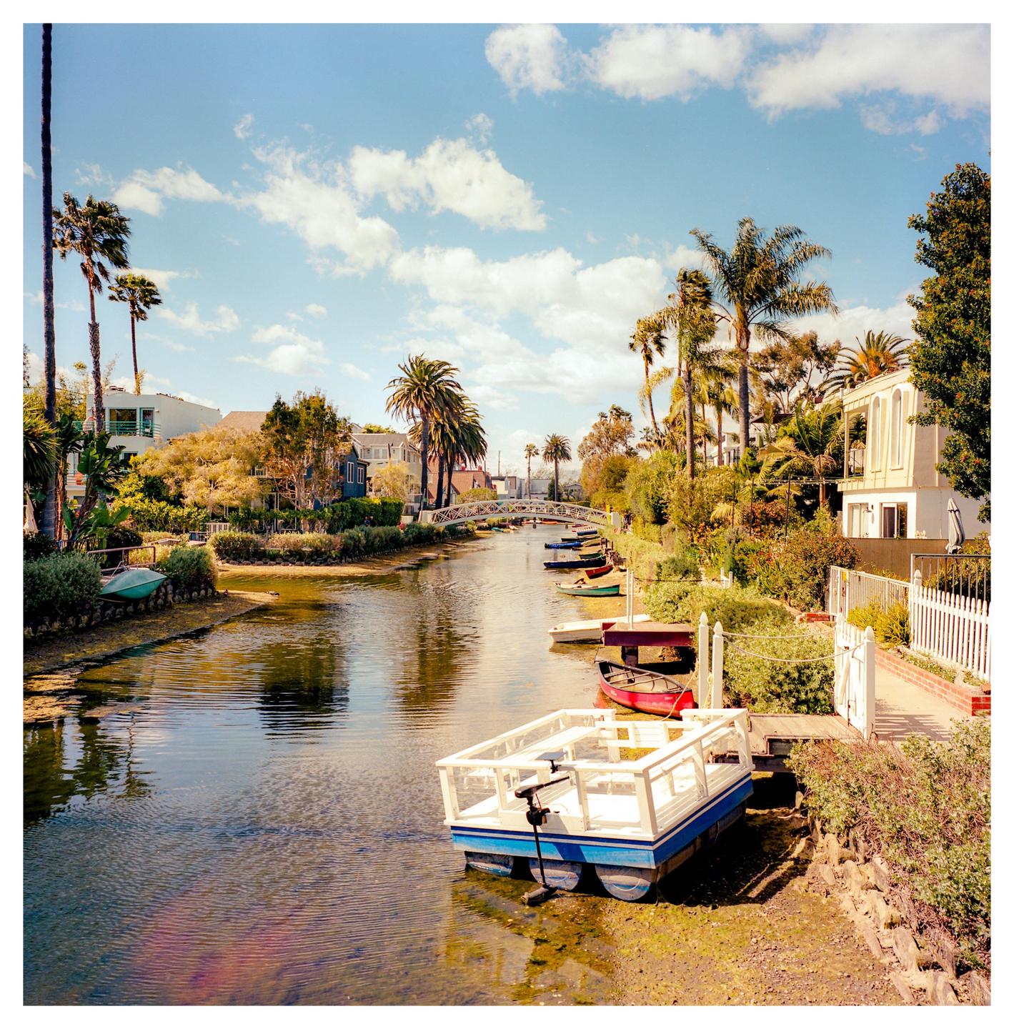 A canal with boats is lined with houses, palm trees, and lush vegetation under a blue sky with clouds.