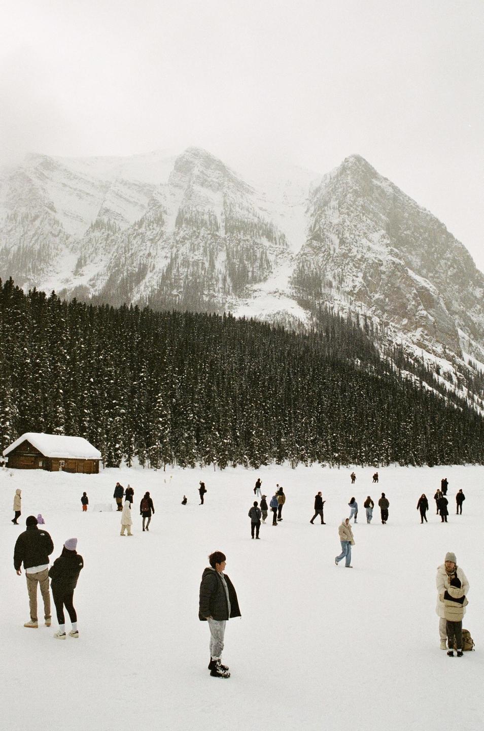 A crowd of people stands on a snow-covered field with a small cabin, a forest, and snow-capped mountains in the background.