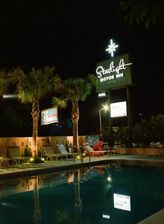 A neon sign for the Starlight Motor Inn stands above a swimming pool and palm trees at night.