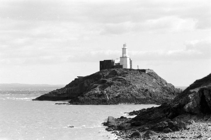 A white lighthouse and building stand on a rocky hill overlooking the sea under a cloudy sky.