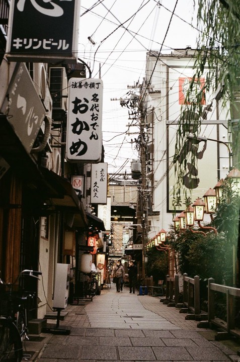 A narrow alleyway in Japan is lined with buildings featuring numerous signs and overhead power lines, with two people walking in the distance.