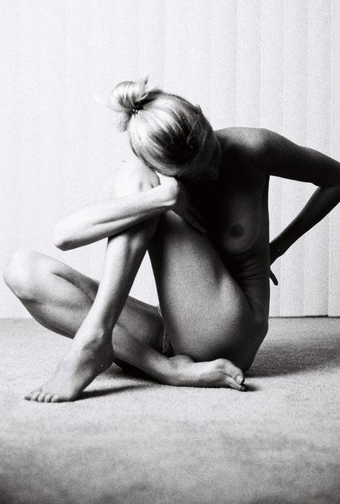 A woman with her hair in a bun sits on the floor with her head bowed and arms crossed over her knees.