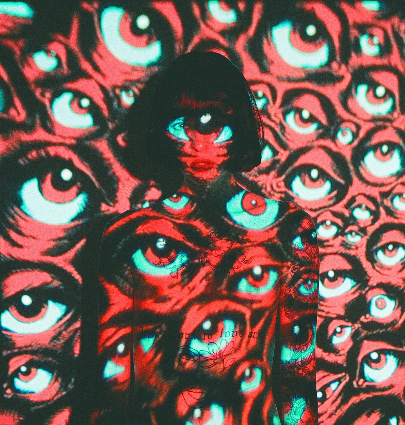 A woman with short dark hair is covered in a projection of many red and blue eyes, with the same pattern on the wall behind her.