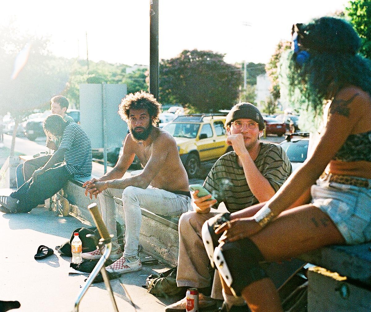 A group of young adults are sitting on a concrete ledge in a park with cars in the background.