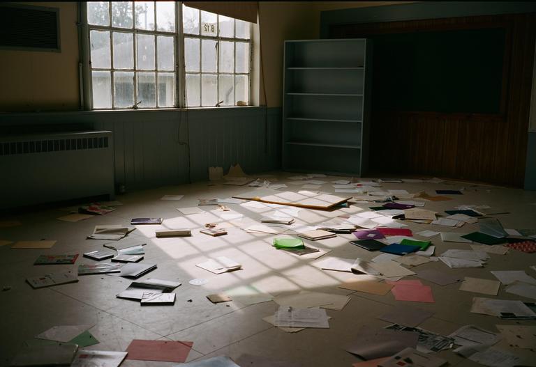 Sunlight streams through a large window into an abandoned classroom with papers scattered across the floor.