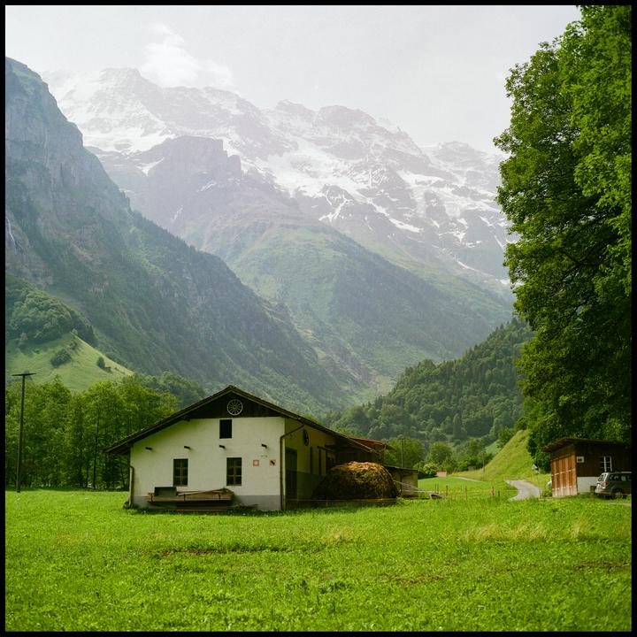 A white house with a hay bale sits in a green field with snow-capped mountains in the background.