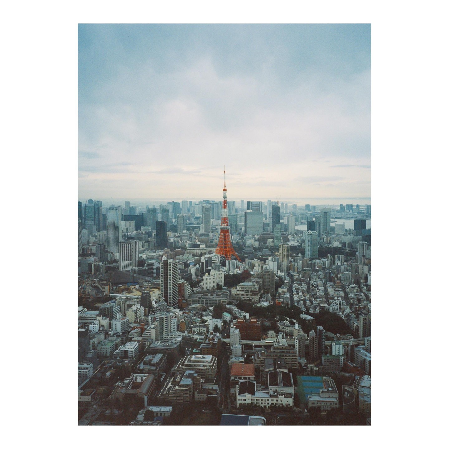 An aerial view shows the Tokyo Tower surrounded by a dense cityscape under a cloudy sky.