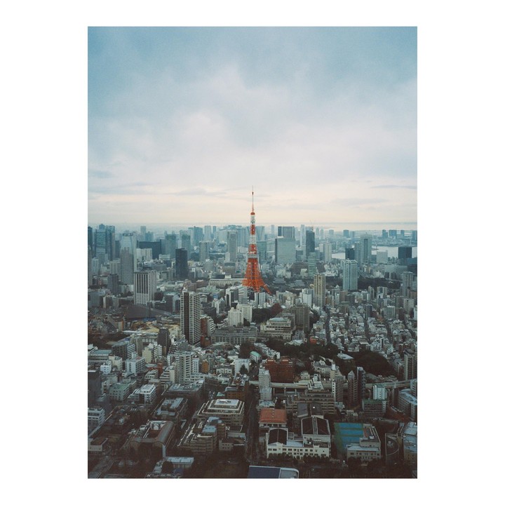 An aerial view shows the Tokyo Tower surrounded by a dense cityscape under a cloudy sky.