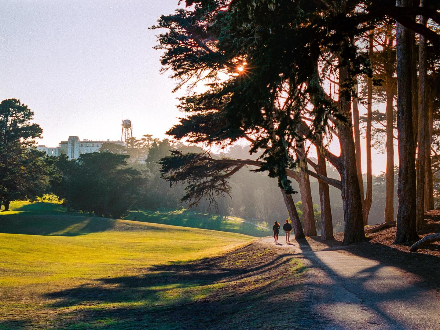 Two people walk on a path next to a golf course with trees and a building in the background.