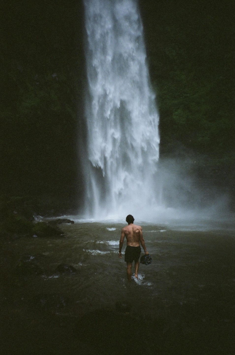 A man walks into a large waterfall, carrying a camera.