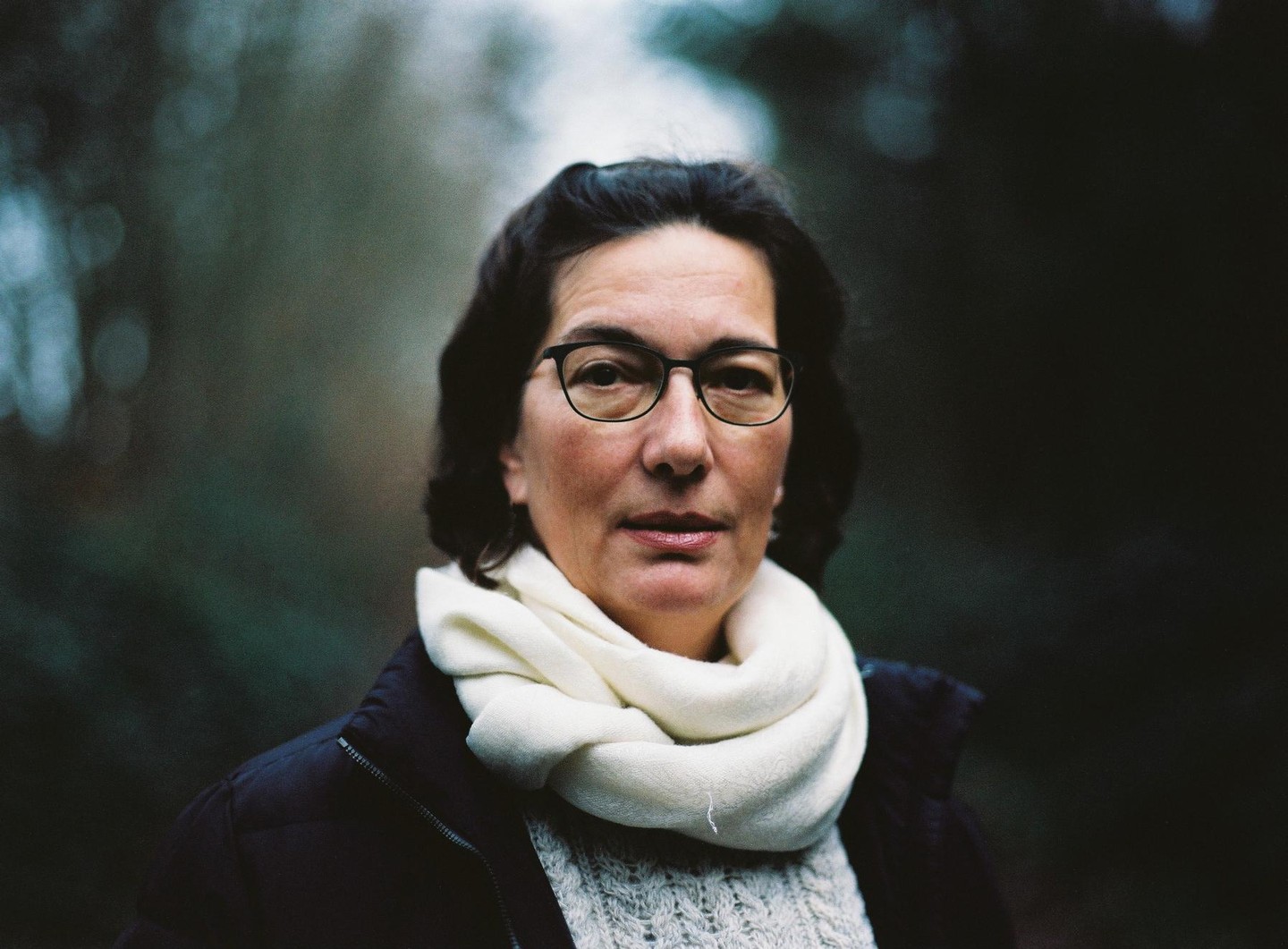 A woman wearing glasses and a white scarf looks directly at the viewer.