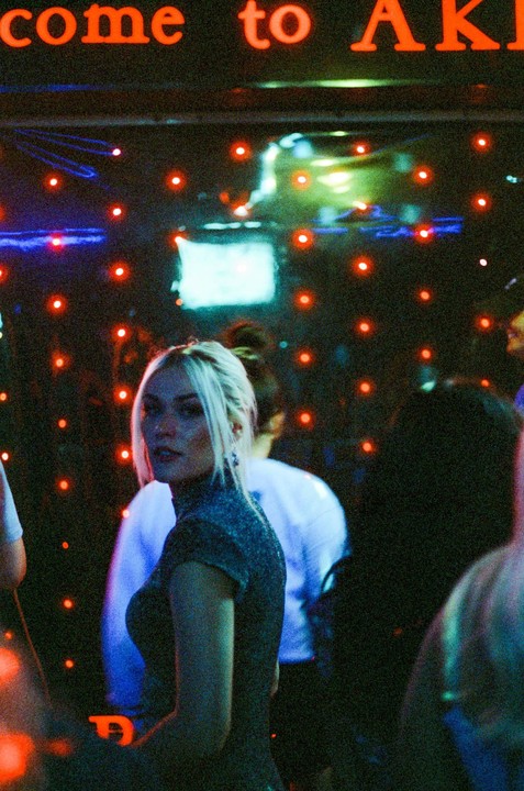 A woman with blonde hair looks over her shoulder in a dimly lit room with red and blue lights.