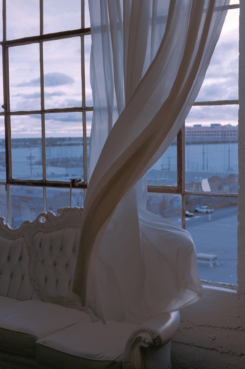 A white curtain blows in front of a large window with a view of a body of water and buildings.