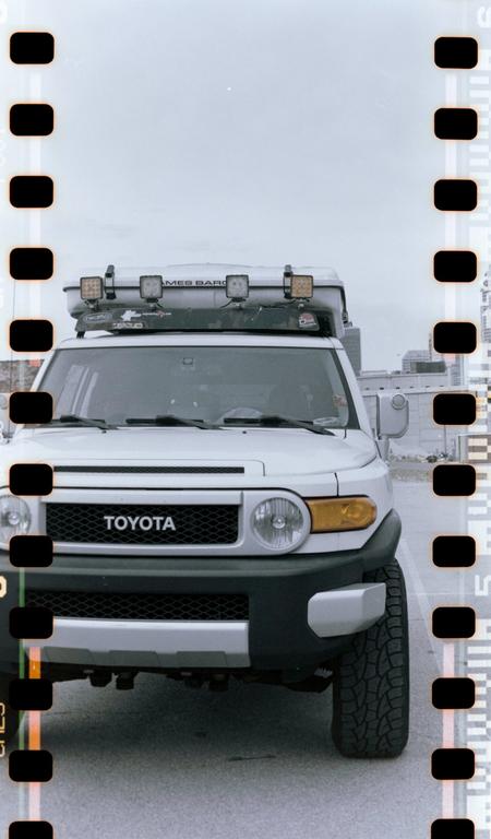 A white Toyota FJ Cruiser with a roof-mounted cargo box and auxiliary lights is parked on an asphalt surface.