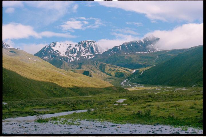A winding river flows through a green valley with snow-capped mountains under a blue sky with clouds.