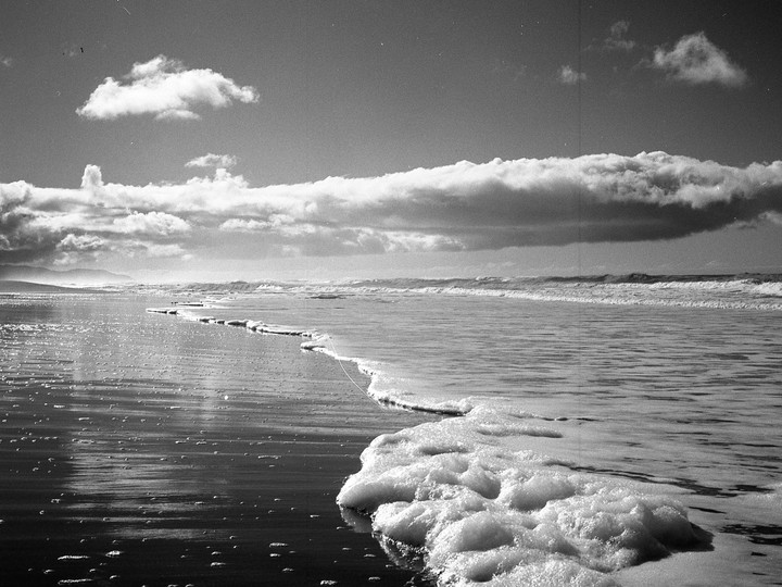 Sea foam covers the wet sand of a beach with waves breaking in the distance under a cloudy sky.
