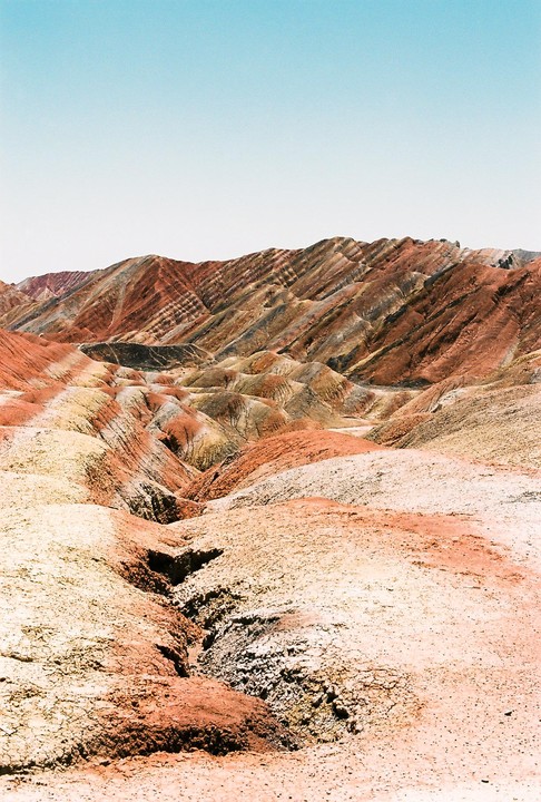 A vast landscape of colorful, striped mountains and dry, cracked earth under a clear sky.