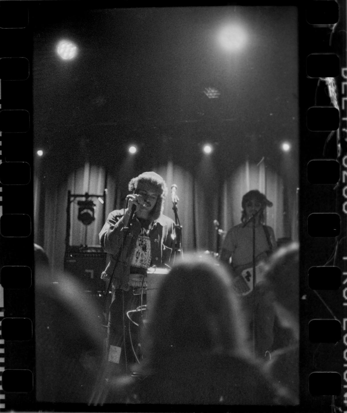 A man sings into a microphone on stage while another man plays guitar in the background.
