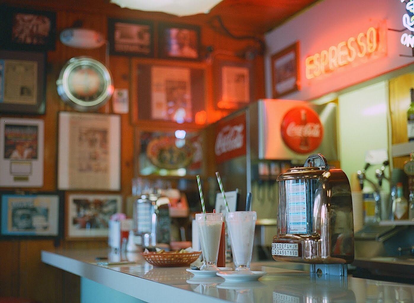 Two milkshakes and a tabletop jukebox sit on a diner counter with a neon "Espresso" sign and Coca-Cola branding in the background.