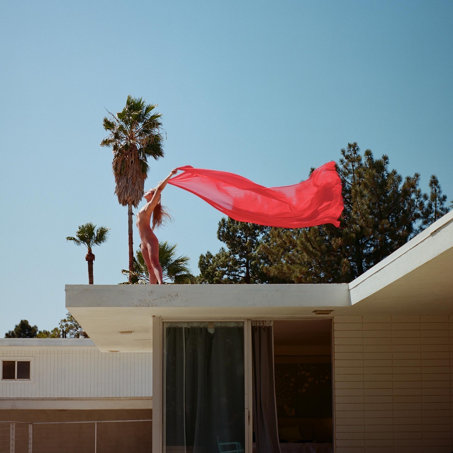 A woman stands on a rooftop holding a red fabric that blows in the wind, with palm trees and a clear sky in the background.