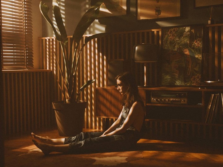 A woman sits on the floor in a room with wooden blinds and a large plant, illuminated by warm, striped light.