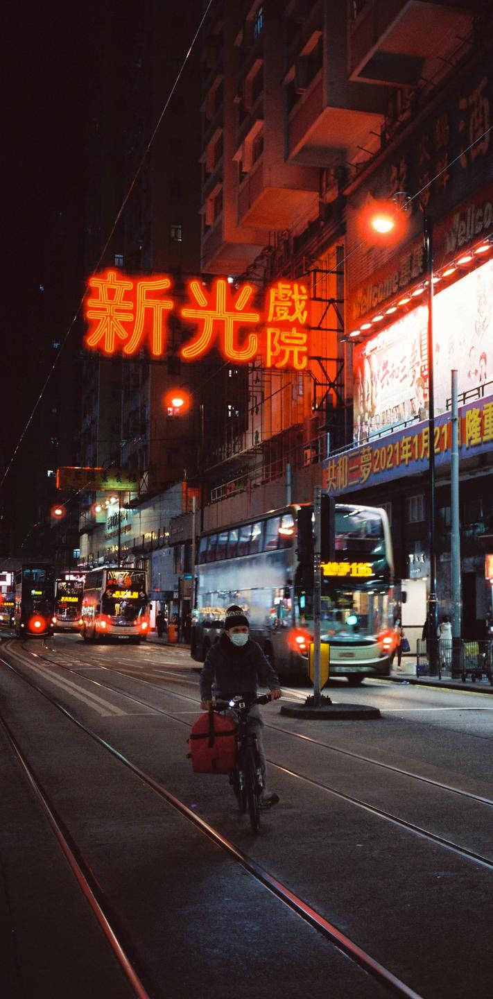 A man on a bicycle with a red delivery bag rides down a city street at night, with neon signs and buses in the background.