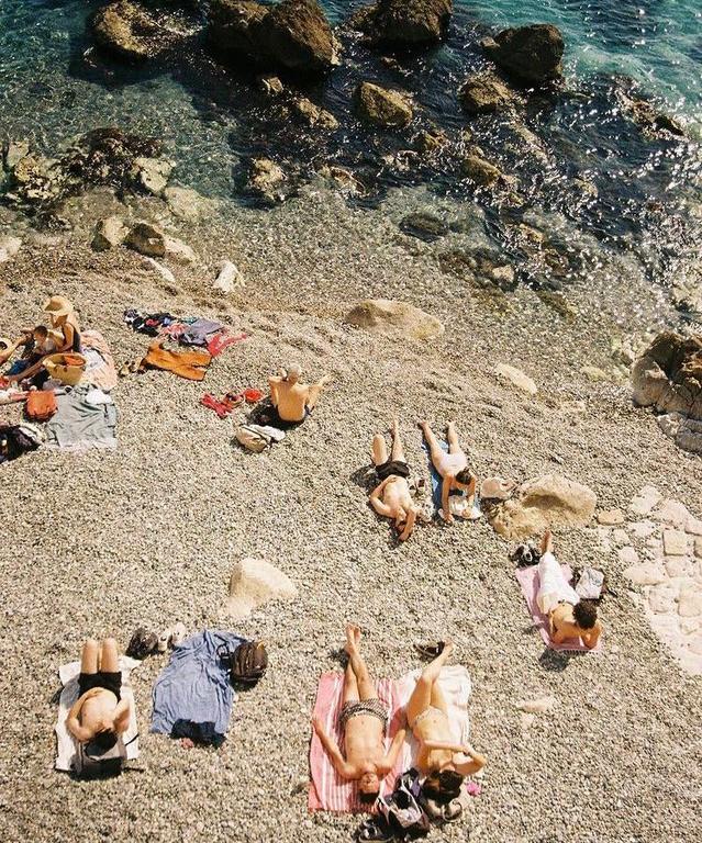 Several people are sunbathing on a rocky beach next to clear blue water.