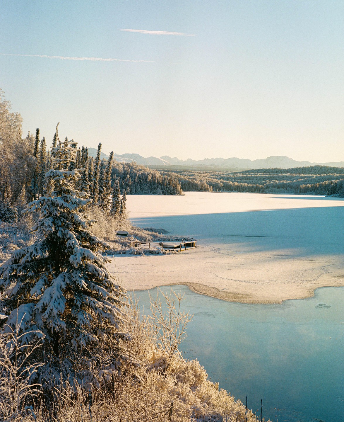 A snow-covered forest and frozen lake are illuminated by golden light under a clear sky.