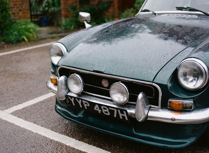 A dark green vintage car with raindrops on its hood is parked on an asphalt surface.