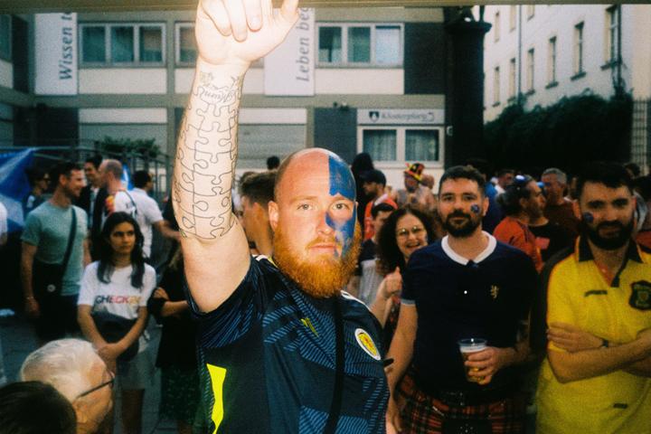 A man with a red beard and blue face paint raises his arm in a crowd of people.