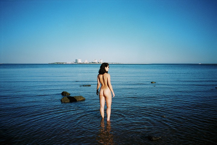 A nude woman stands in the ocean with her back to the viewer, looking towards a distant city skyline.
