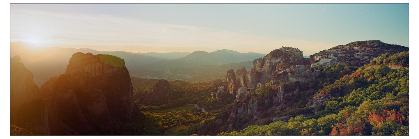 A panoramic landscape shows monasteries built on rock formations in Meteora, Greece, at sunset.