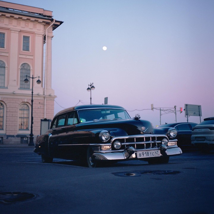 A black vintage Cadillac is parked on a street with a full moon visible in the sky.