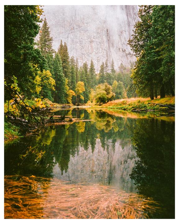 A river reflects a forest and a large rock face under a hazy sky.