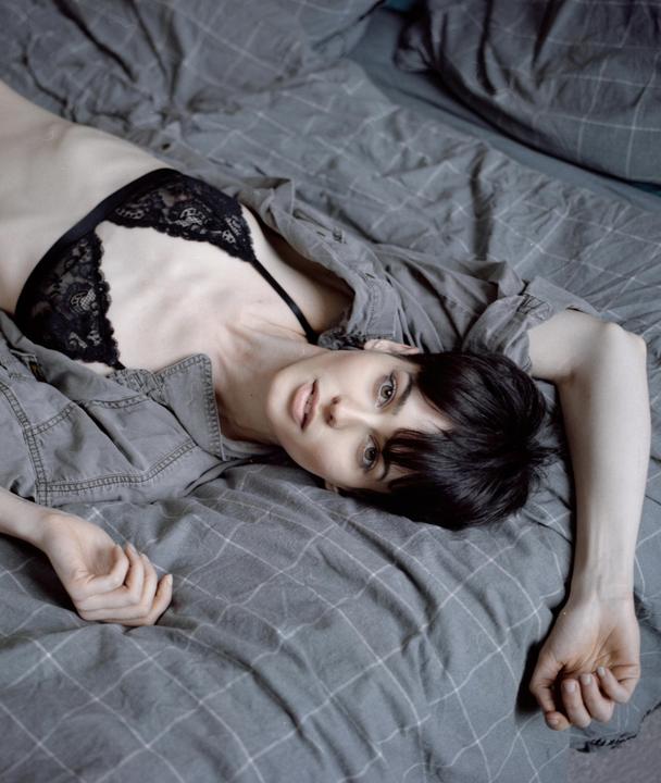 A woman with short dark hair and a black lace bra lies on a bed with gray sheets and a gray shirt.